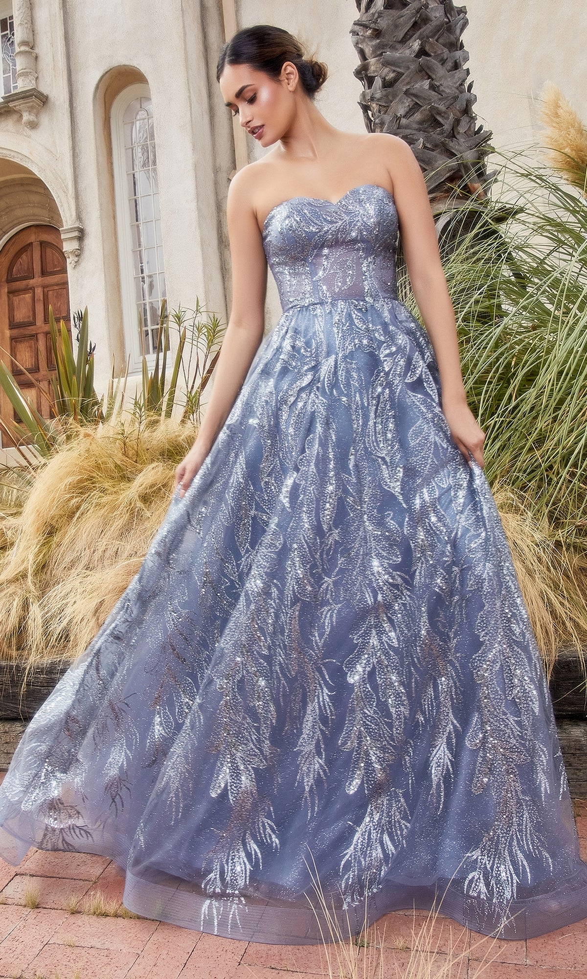 A woman wearing the Strapless Long Prom Ball Gown with Capelet A1174 stands outdoors near tall grasses and a palm tree. The elegant silver-blue gown adds sophistication to the tranquil setting.